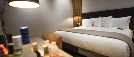 A hotel room with a neatly made bed, a bedside table with toiletries, a lamp, and a wooden panel wall.