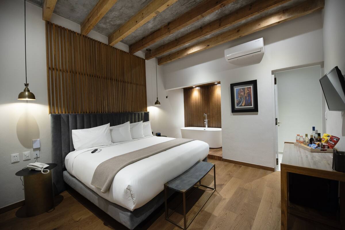 Habitació d'hotel moderna amb un llit gran, una taula de nit i una banca. Hi ha un bany amb banyera i plat de dutxa.