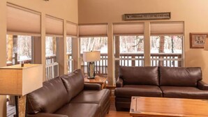 A spacious living room with two leather sofas, a wooden coffee table, and a ceiling fan. Large windows provide natural light and a view of the outdoors. - Mt. Shavano Chalet at Creekside chalets (Salida)