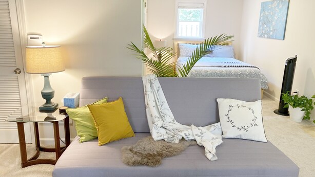 Une chambre avec un canapé gris, une table de chevet avec une lampe, un lit recouvert d’une couvre-lit bleue et blanche, une fenêtre et un tableau accroché au mur.