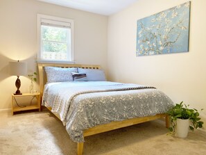 A bedroom with a wooden bed, a bedside table with a lamp, a window, and a wall-mounted painting. - Cozy Urban Retreat - Entire Guest Suites in Buckhead (Atlanta)
