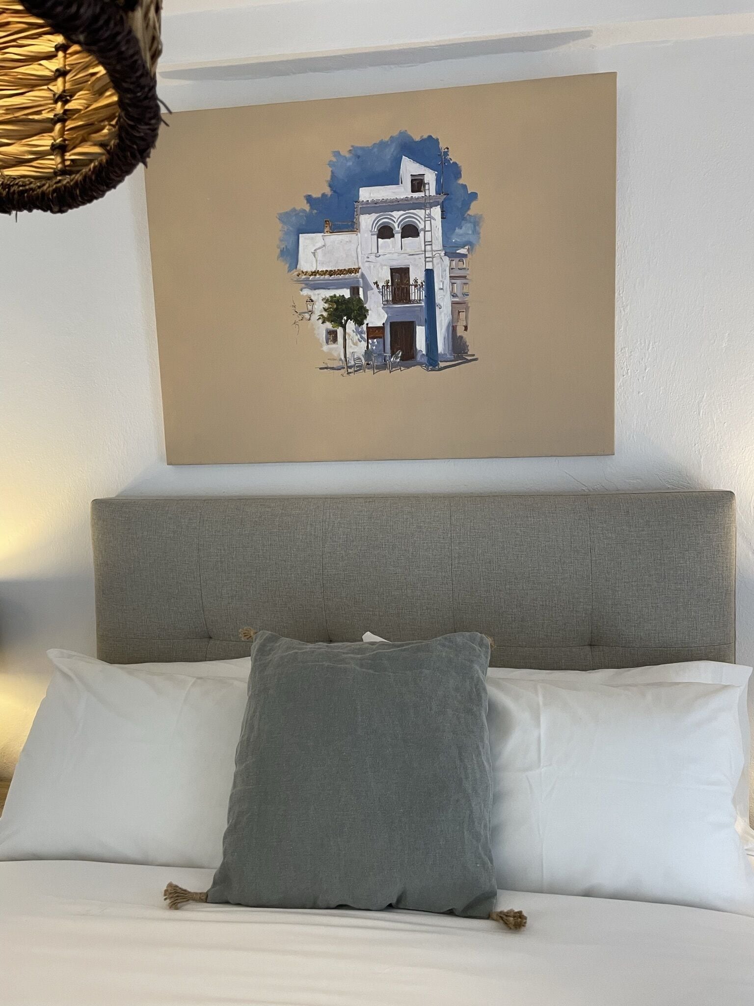 A bedroom with a bed, a grey headboard, and a wall-mounted painting of a building.