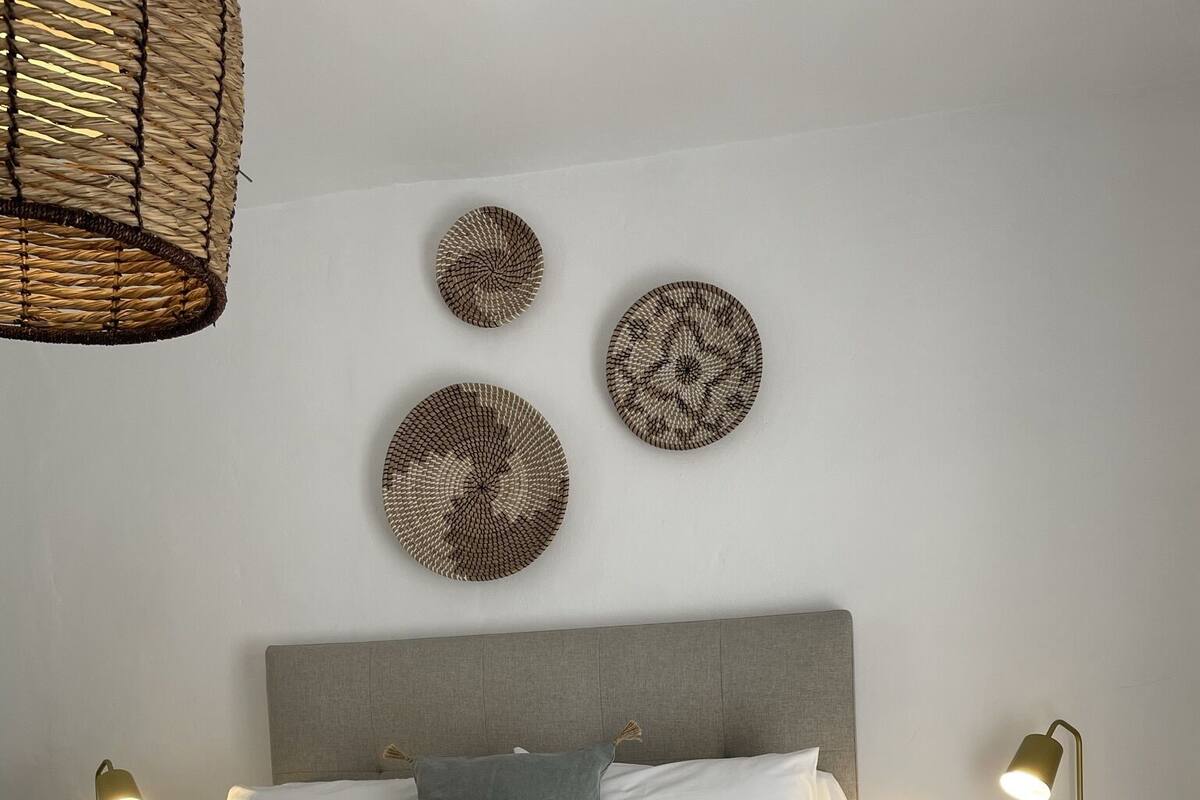 Una cama bien tendida con ropa de cama blanca y gris, un plaid gris y una toalla enrollada con un adorno. Hay dos mesitas de noche con lámparas, una obra de arte circular tejida en la pared y un cabecero gris.