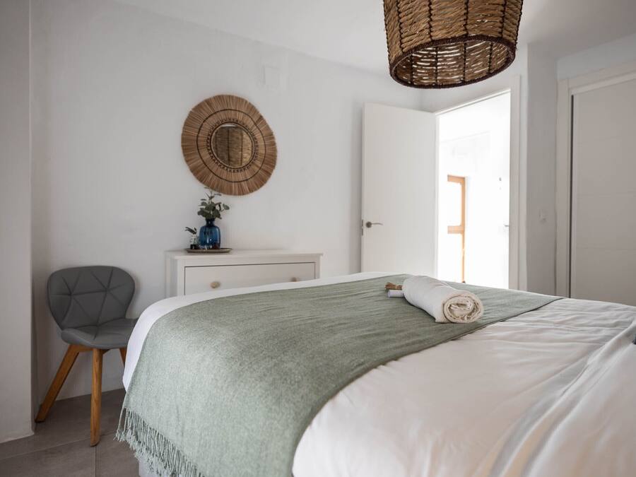 Un dormitorio ordenado con cama, una silla, cómoda y lámpara de fibras naturales.