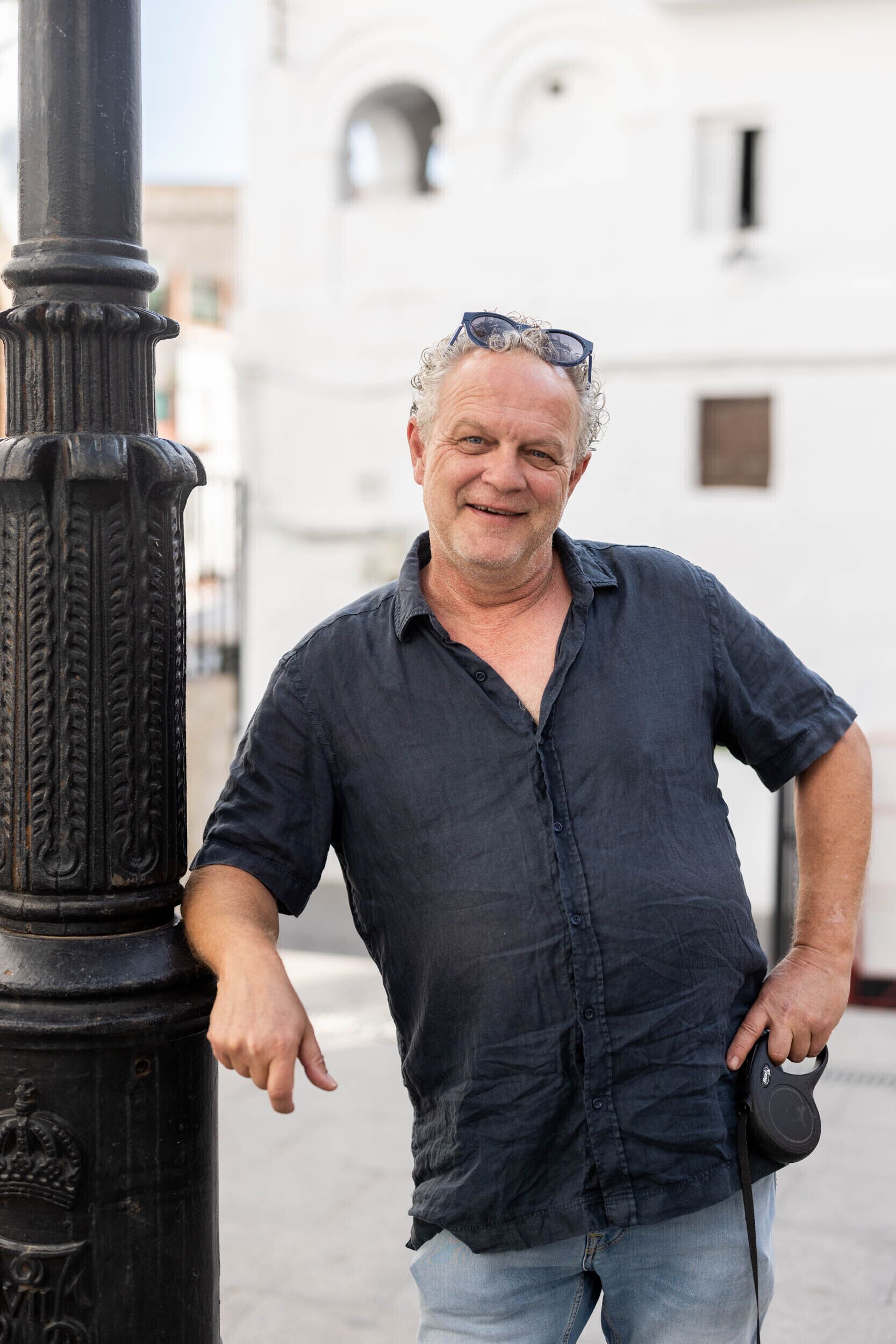 A man standing next to a black pole with a crown emblem, wearing a dark shirt and holding a camera.