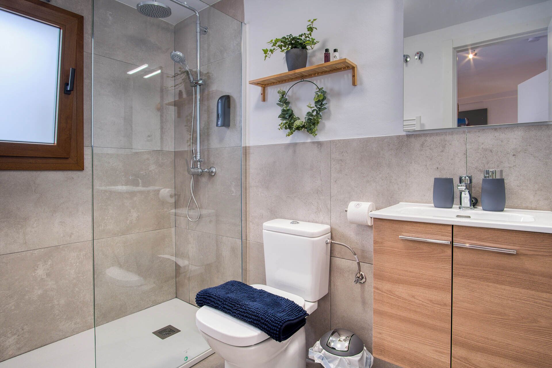 A modern bathroom with a glass-enclosed shower, a toilet with a blue towel, a wooden vanity with a mirror, and a shelf with toiletries.