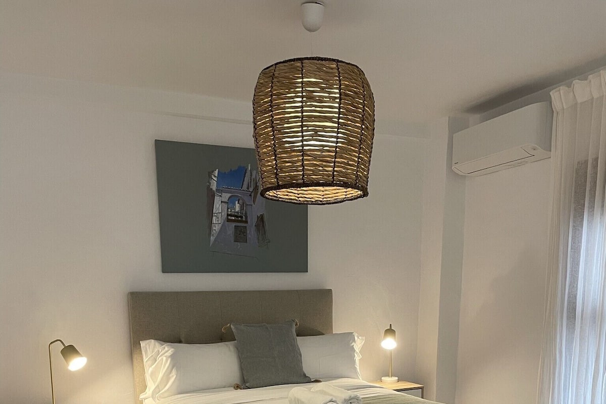 Un dormitorio moderno con cama, mesitas de noche, una lámpara colgante y una pequeña mesa redonda con una planta.