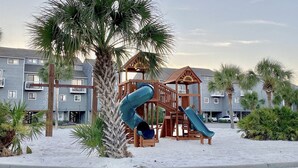 Un parque infantil con toboganes y estructuras para escalar, rodeado de palmeras y un edificio con balcones al fondo.
