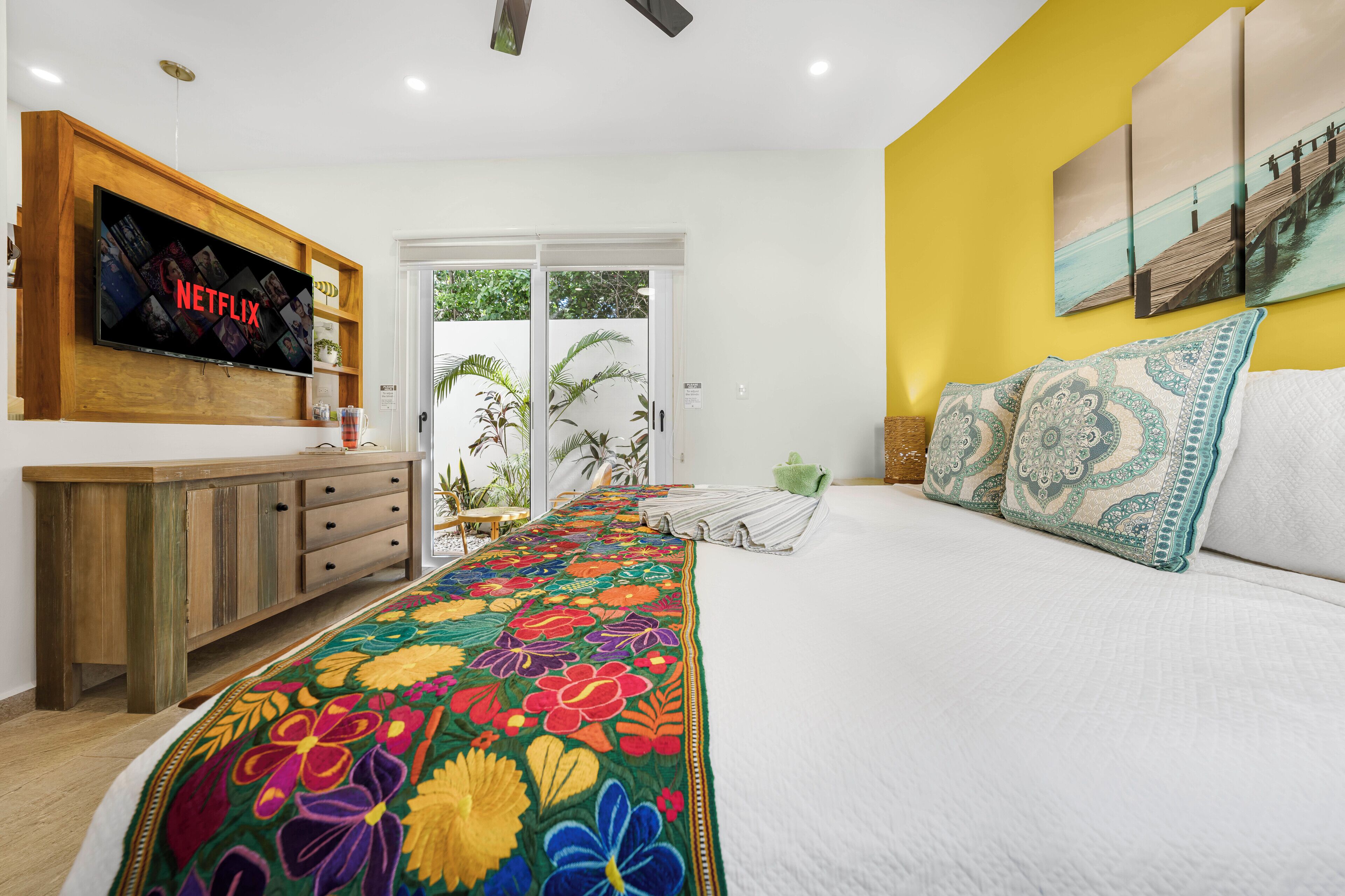 A bedroom with a large bed, a TV mounted on a wooden cabinet, a view of the outdoors through a sliding door, and a yellow accent wall.
