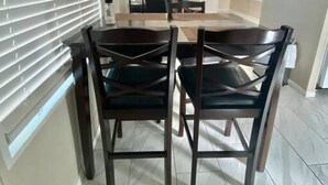 A dining area with a round table and four chairs, a large window with blinds, and a view of a room with a bed and a chair.