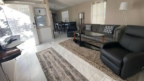 A living room with a sofa, a coffee table, and a TV. There is a kitchen area with a fridge and a dining table.