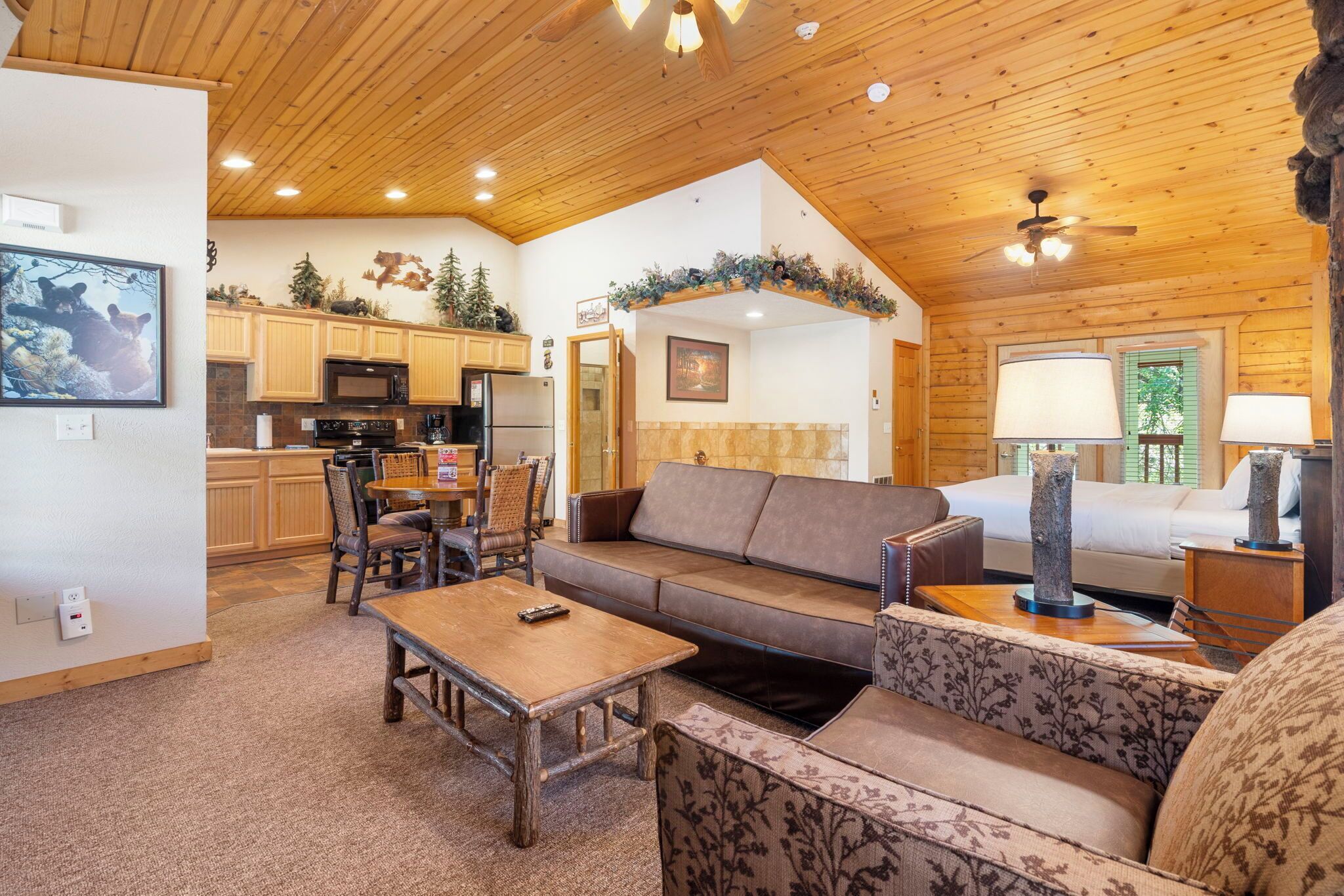 A cozy cabin interior with a wooden ceiling, a dining area with chairs, a sofa, a coffee table, and a kitchen with wooden cabinets and appliances.