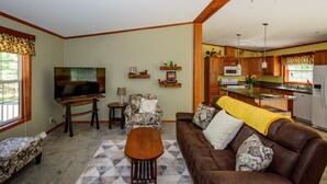 Uma sala de estar com um sofá de couro marrom, uma mesa de centro de madeira e uma televisão. Cozinha visível ao fundo, com armários de madeira e um micro-ondas.