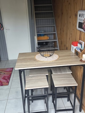 Un tavolo in legno con un sottopiattino intrecciato, due sgabelli neri, un piccolo cestino in filo nero e un cartello con la password del WiFi.