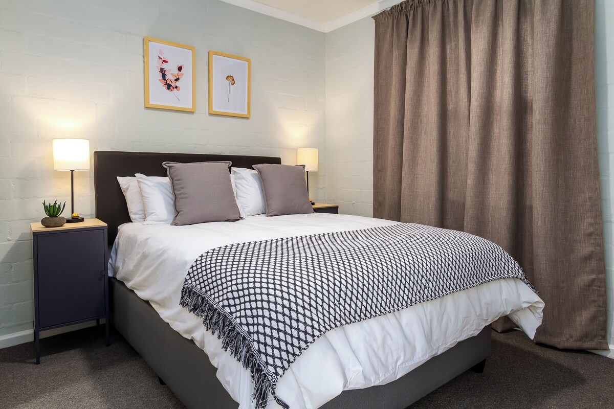 A neatly made bed with a patterned throw, two bedside lamps, and a small plant on a nightstand.