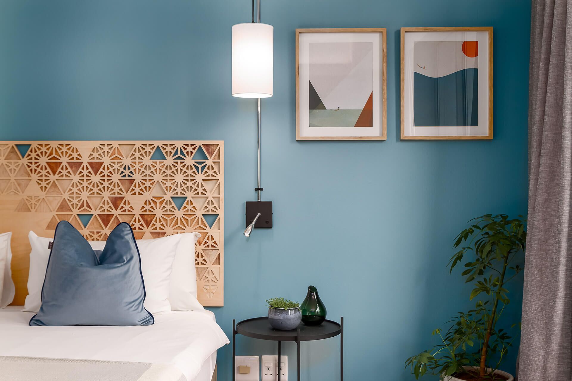 Ein Schlafzimmer mit einem Kopfende im geometrischen Muster, einem Bett mit blauen und weißen Kissen, zwei gerahmten abstrakten Kunstwerken, einem kleinen runden Tisch mit einer Pflanze und einer weiteren Pflanze neben dem Bett.