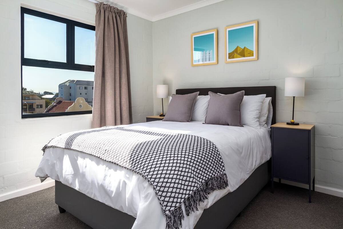 A bedroom with a bed, two bedside tables, a window with curtains, and two framed pictures on the wall.