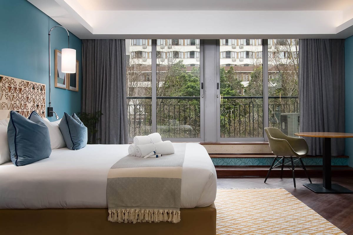 A hotel room with a large bed, a desk, and a view of buildings through the windows.