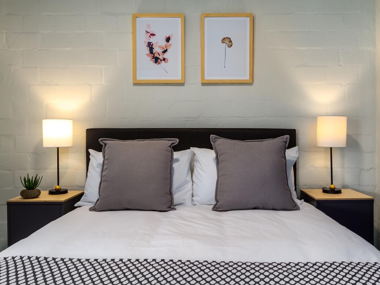 Un lit bien fait, avec deux oreillers, un plaid à motifs et deux tables de chevet avec des lampes. Deux œuvres d’art encadrées sont accrochées au mur au-dessus du lit.
