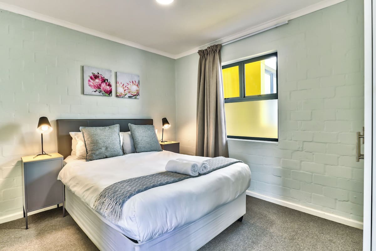 A neatly made bed with a grey and white duvet, two bedside lamps, and a window with curtains.
