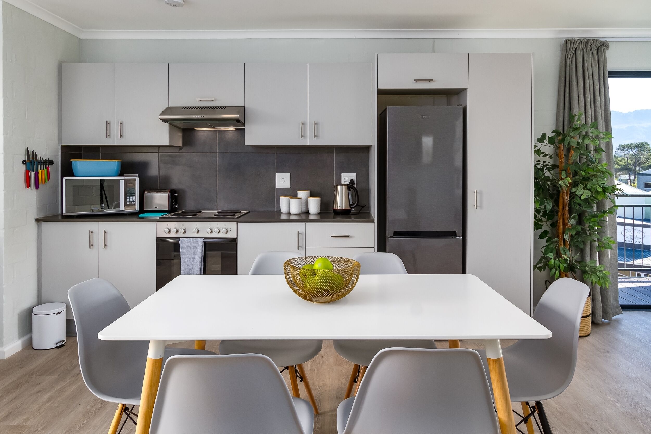 A modern kitchen with white cabinets, a microwave, stove, and a refrigerator. There is a dining table with chairs and a bowl of fruit.