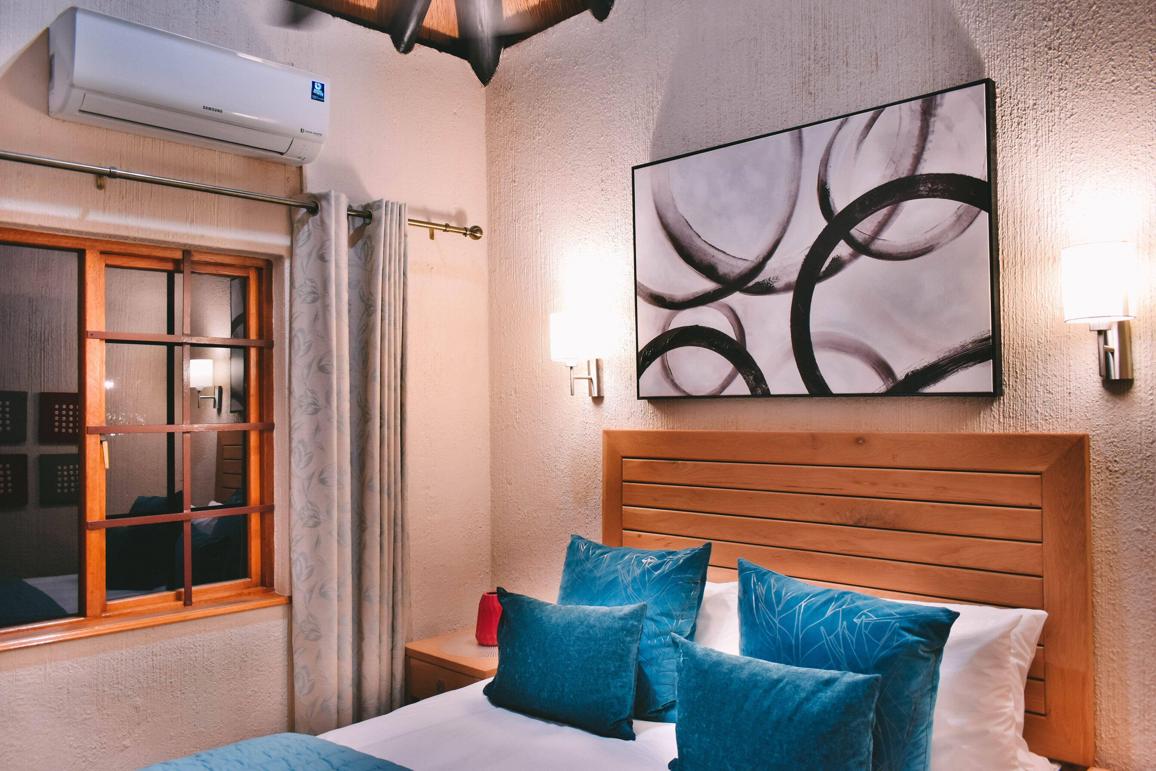 Habitación de hotel con cabecera de madera, un cuadro abstracto grande, una ventana con cortinas y un equipo de aire acondicionado.