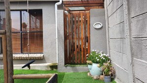 Uma casa com um portão de madeira com a placa "NICHOLAS'S GATE", um pequeno jardim com um balanço e um vaso com flores brancas.