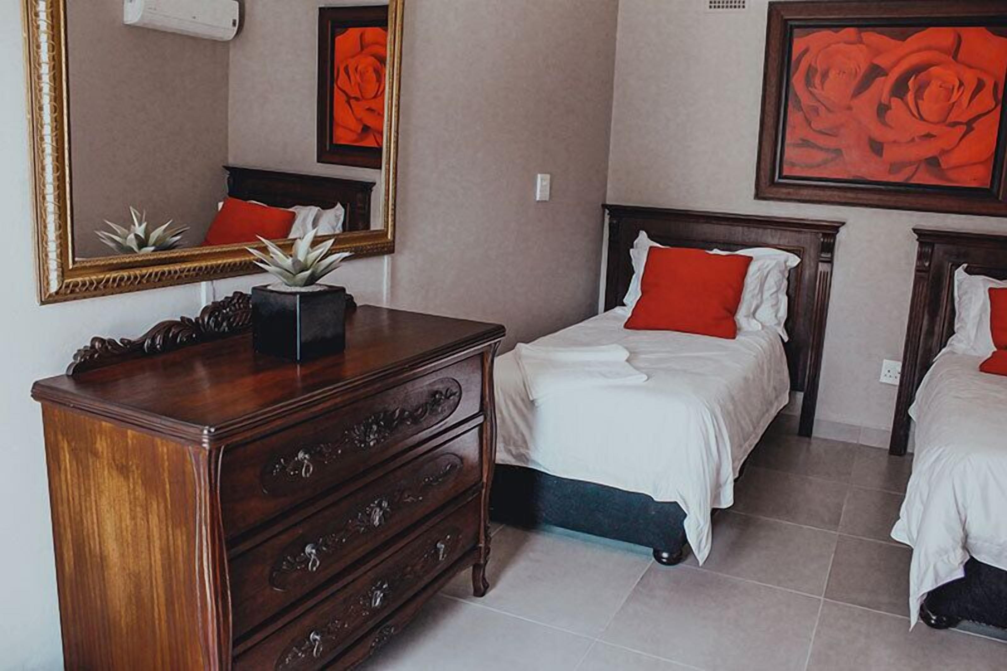A bedroom with a wooden dresser, two beds with red and white bedding, a large mirror, and a framed painting of a rose.