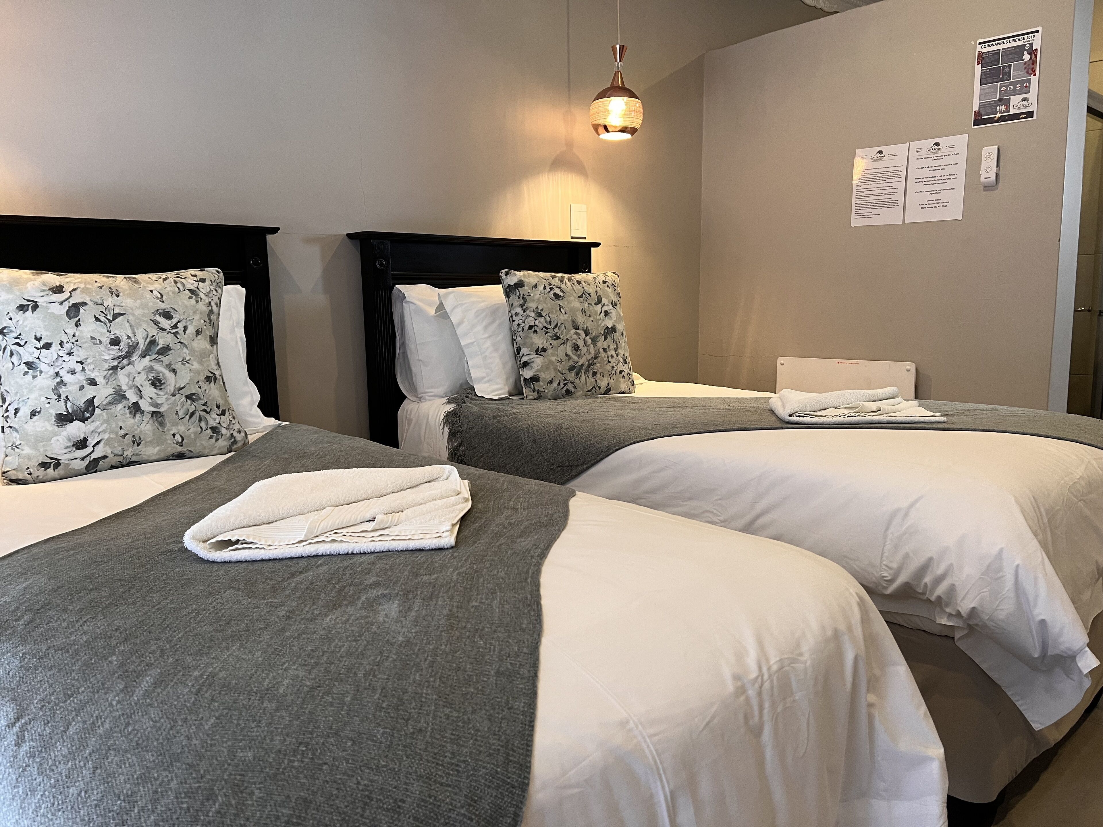 Ett hotellrum med två sängar, var och en med ett grått och vitt täcke, ett tr äfärgat sänggavel och en enkel hängande lampa.