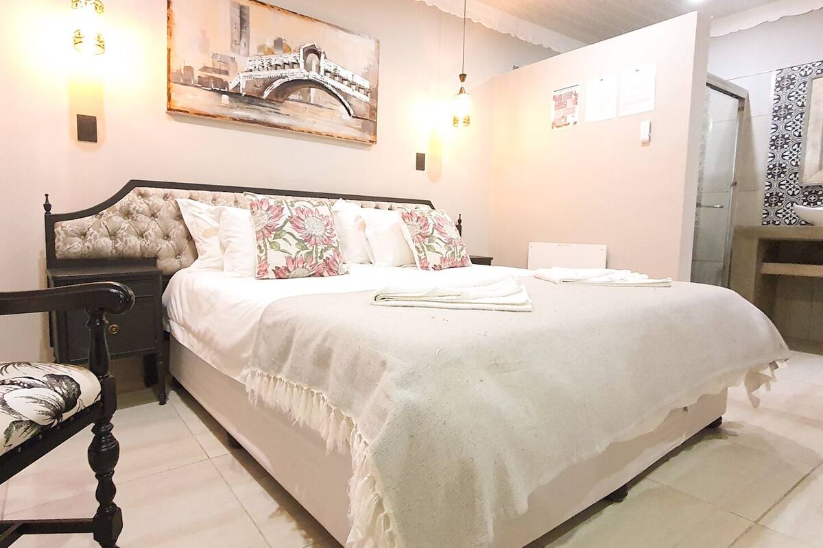 A bedroom with a bed, a nightstand, a chair, and a bathroom with a sink and mirror.