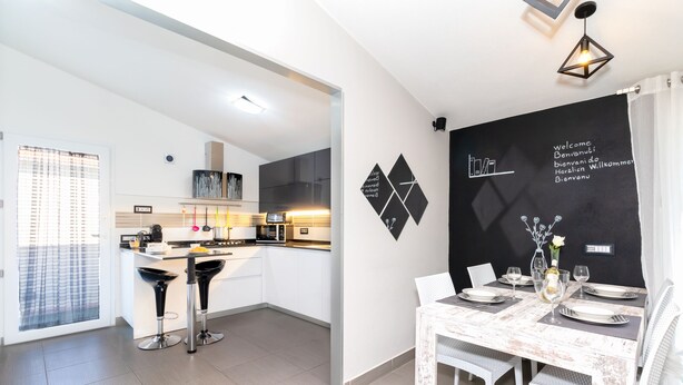 A modern kitchen with white cabinets, a dining area with a white table and chairs, and a blackboard with multilingual welcome messages. - Sunset Bay Tecno Home Sardinia (Valledoria)