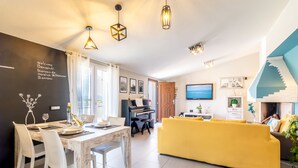 A modern living room with a piano, yellow sofa, and a dining area with white chairs. - Sunset Bay Tecno Home Sardinia (Valledoria)