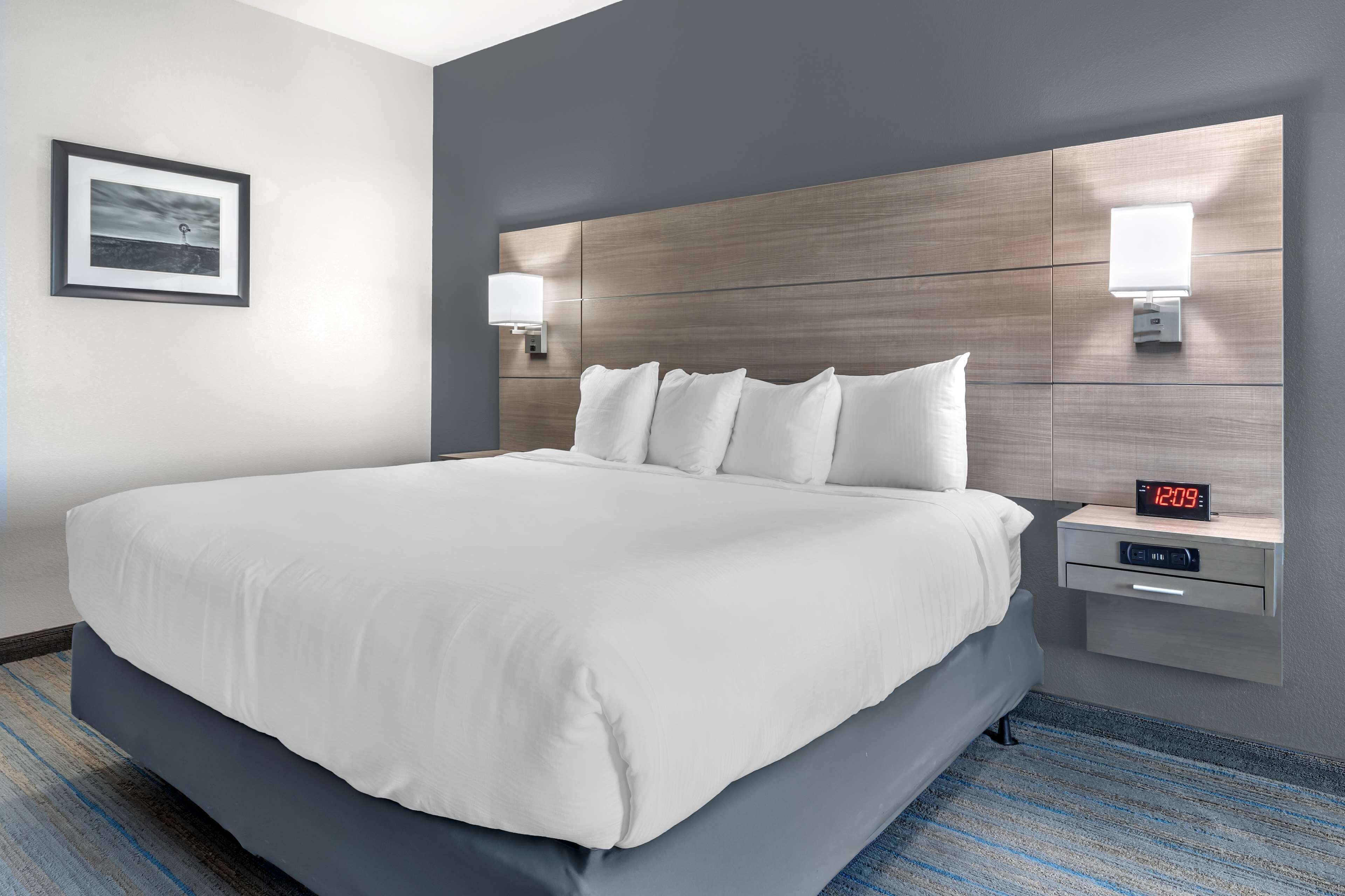 Una cama bien tendida con sábanas blancas, cabecera de madera, lámparas de noche, un reloj digital y un cuadro en la pared.