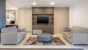A modern living room with two grey sofas, a central ottoman, a flat-screen TV mounted on a wooden wall, and recessed ceiling lights. - Best Western Plus Stephenville Inn (Stephenville)