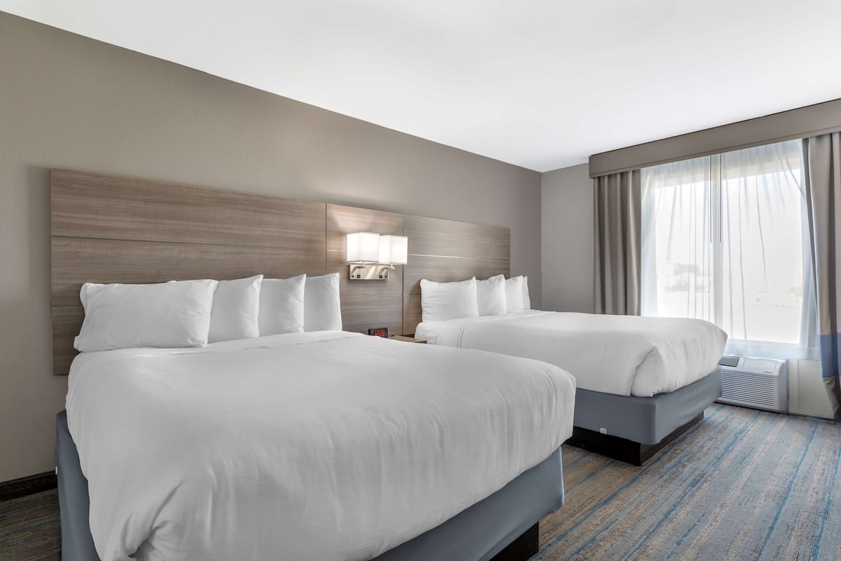 A hotel room with two beds, a large window with curtains, a wall-mounted lamp, and a carpeted floor.