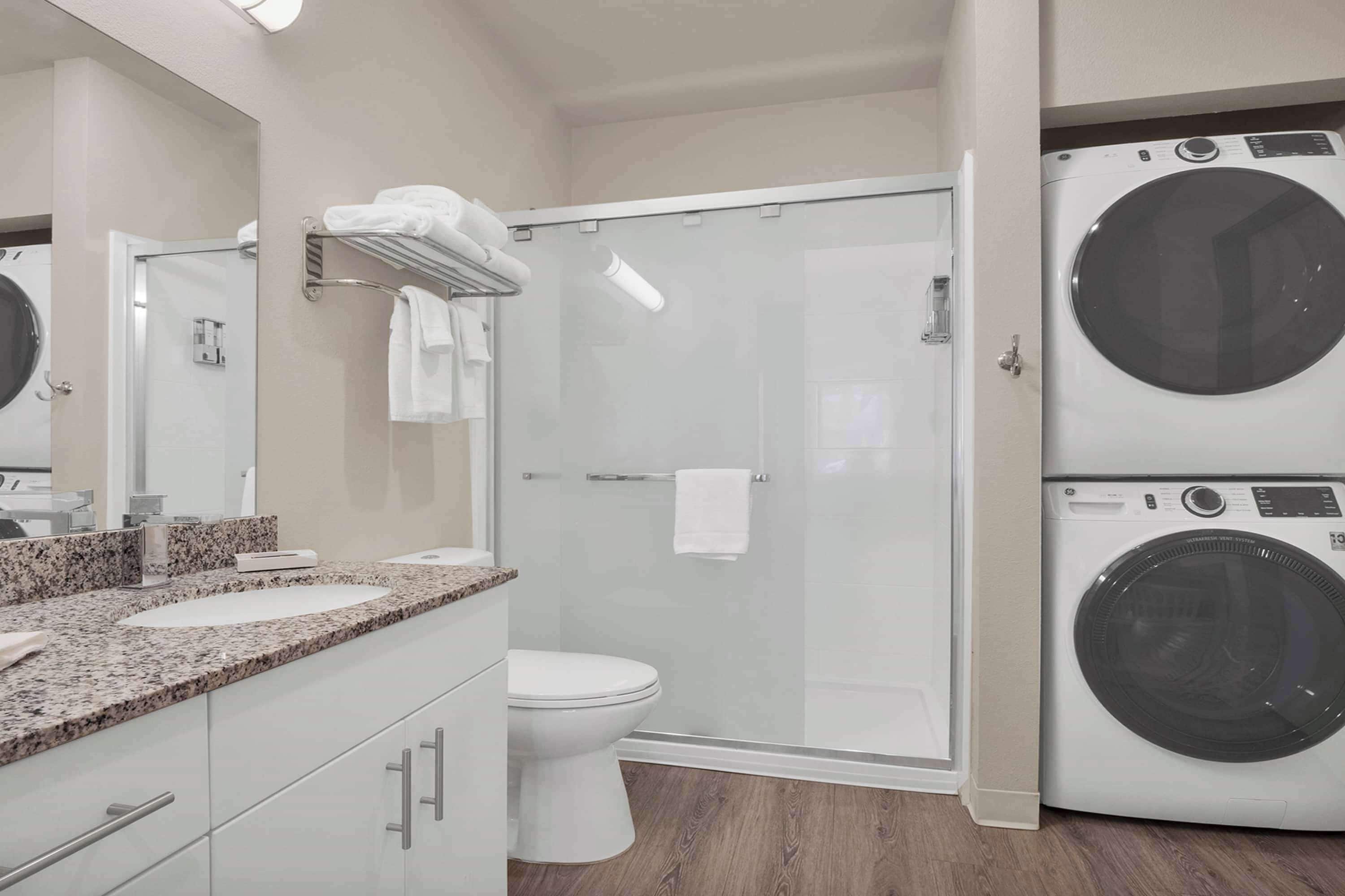 A modern bathroom with a granite countertop, a white toilet, a glass-enclosed shower, and a front-loading washer.