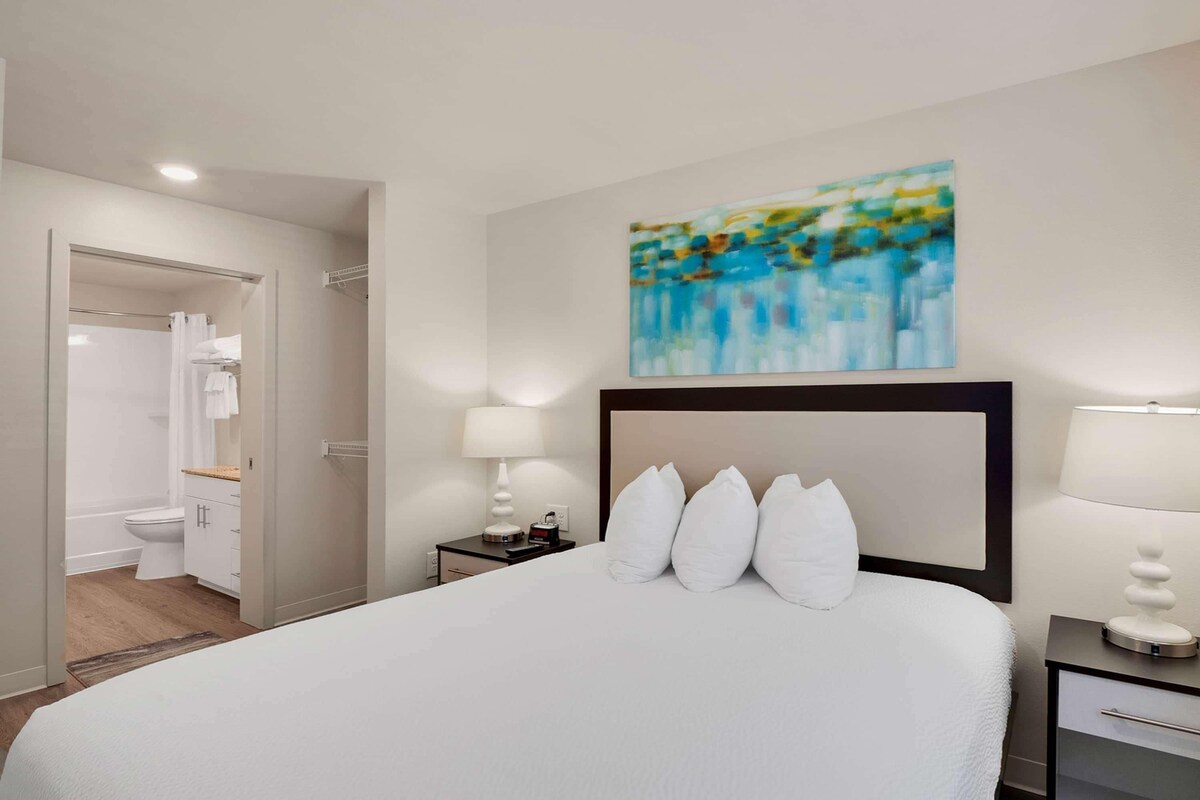 Une chambre d’hôtel avec un grand lit, une table de chevet avec une lampe, un tableau au mur et une salle de bain visible à travers une porte ouverte.