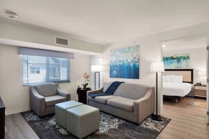 A modern hotel room with a sofa, armchair, and ottoman, a large window with blinds, a painting on the wall, and a bed with a nightstand.