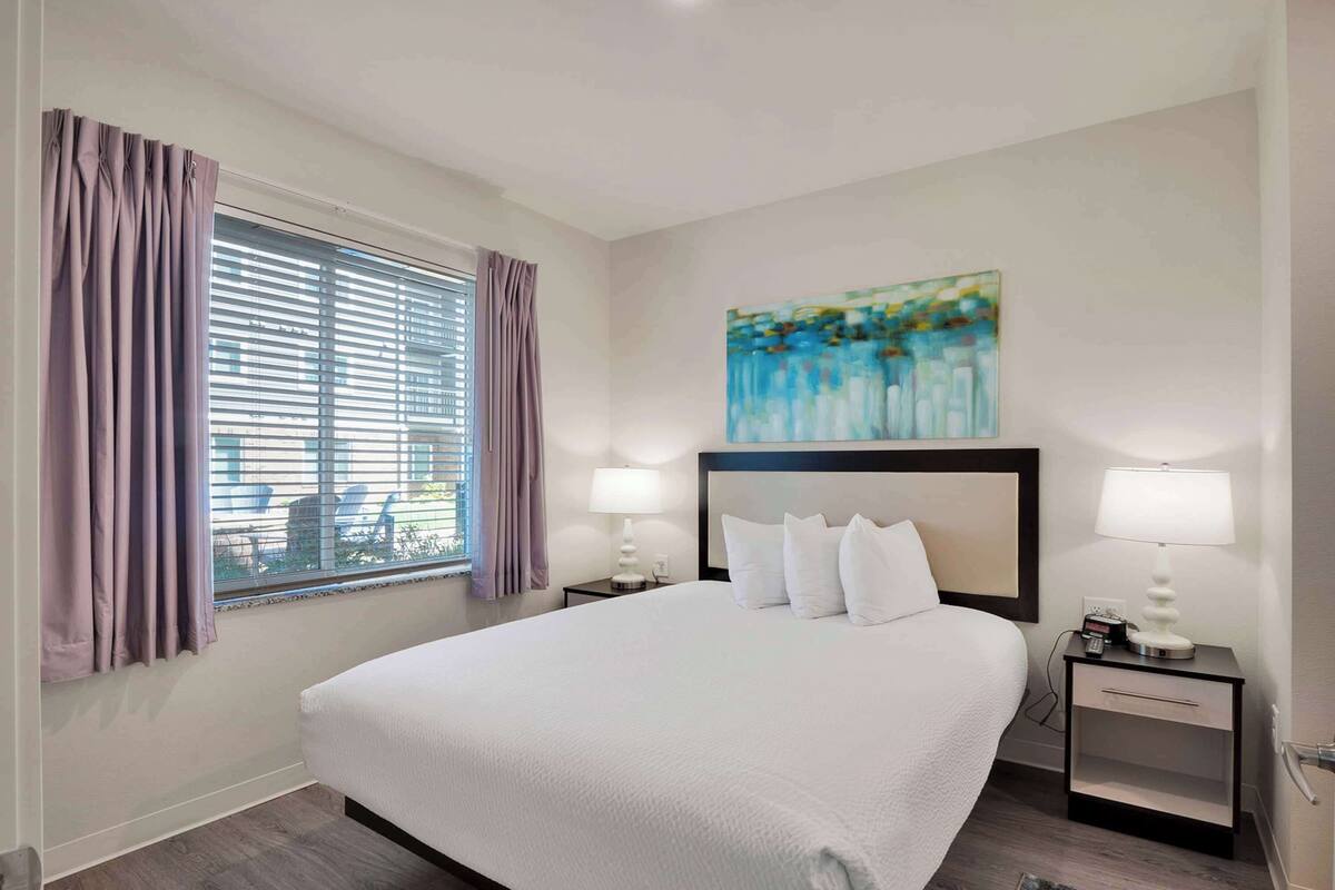 Een hotelkamer met een groot bed, twee nachtkastjes met lampen, een schilderij aan de muur en een raam met gordijnen.