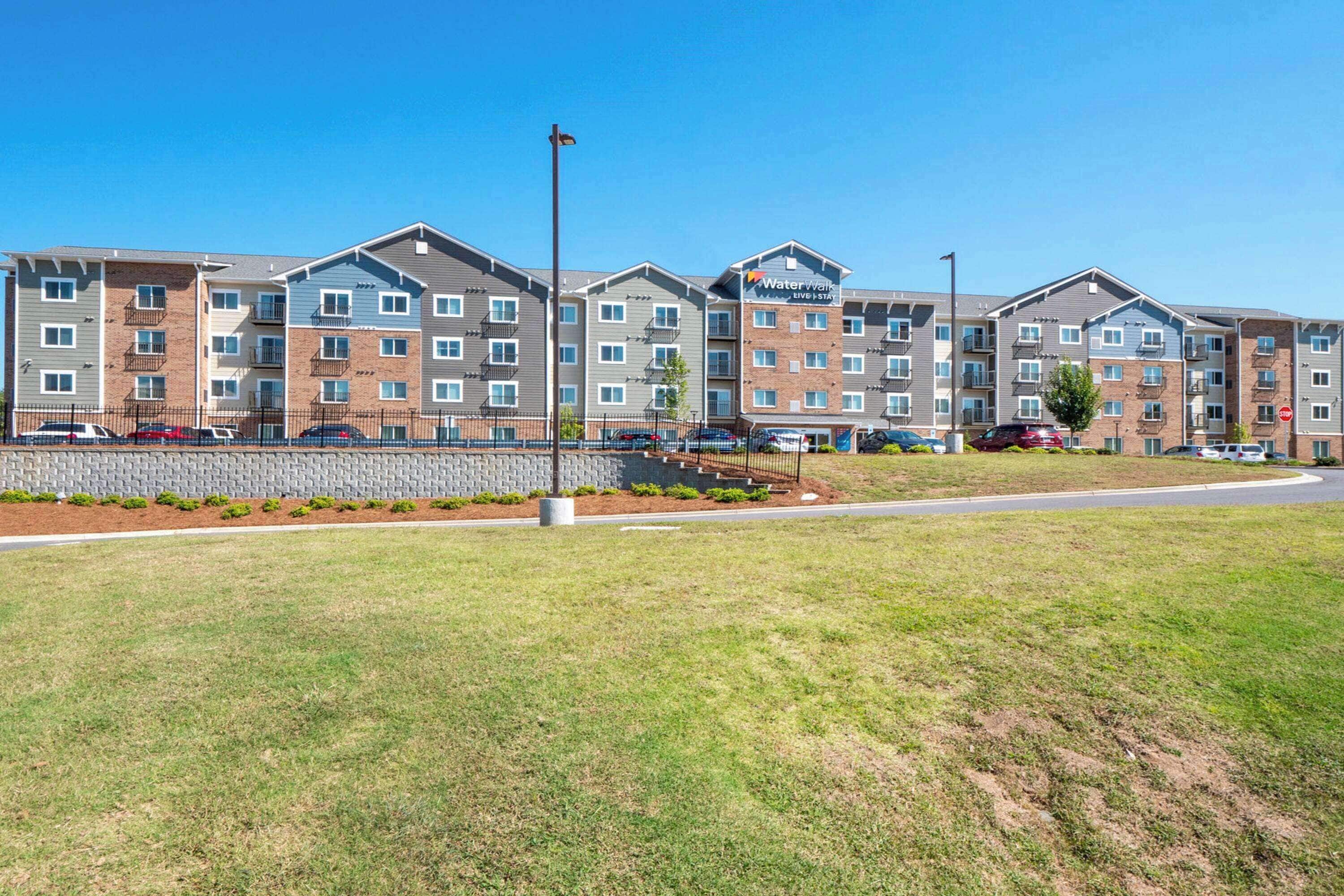 A multi-story apartment building with a brick facade, a landscaped lawn, and a parking area with cars.