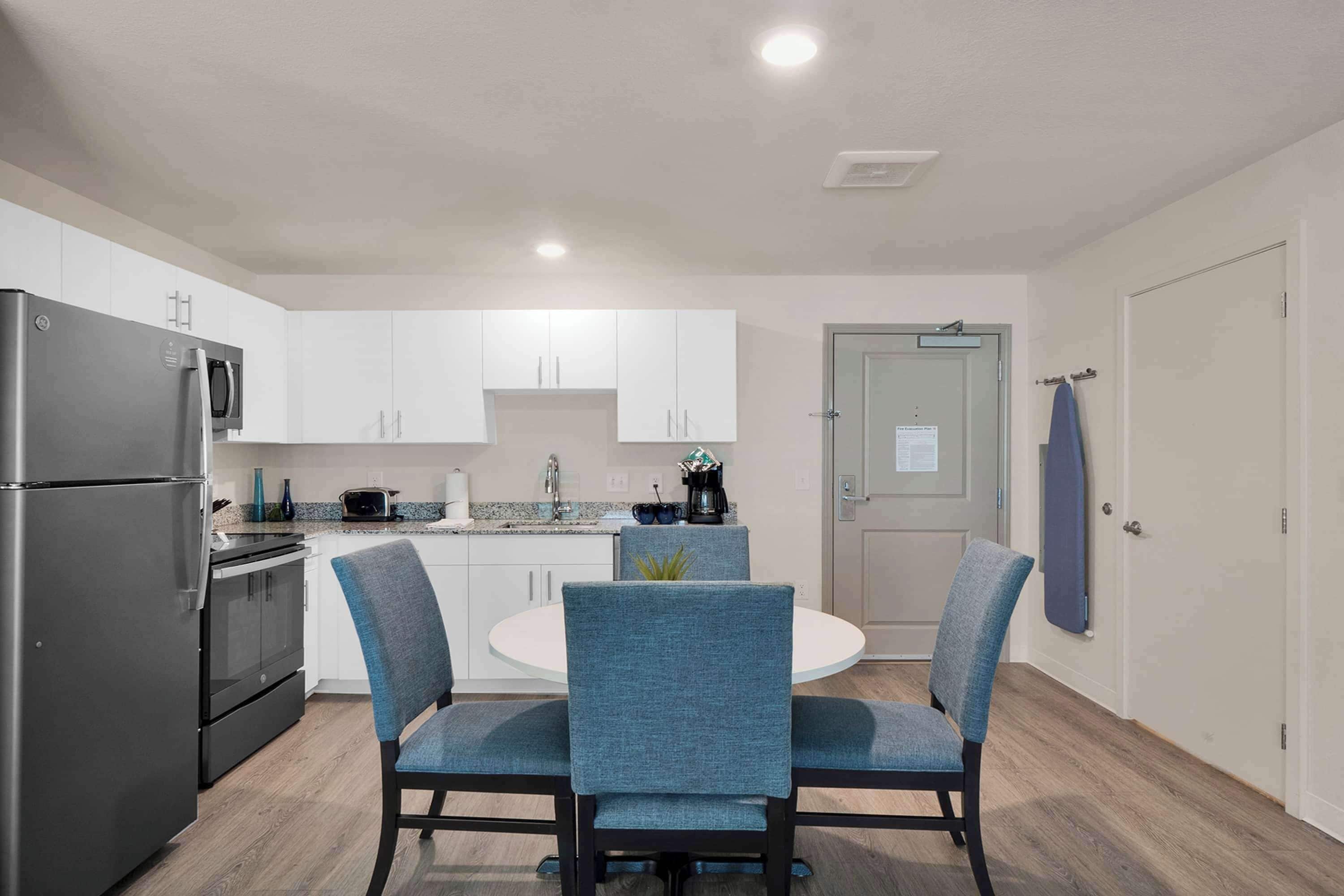 A modern kitchen with white cabinets, a stainless steel refrigerator, a microwave, and a coffee maker. There is a round dining table with blue upholstered chairs.