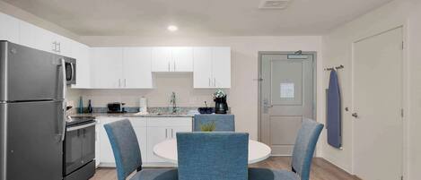 A modern kitchen with white cabinets, a stainless steel refrigerator, a microwave, and a coffee maker. There is a round dining table with blue upholstered chairs.