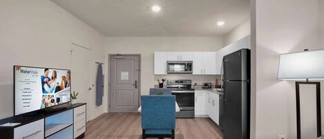 A modern kitchen with white cabinets, a black refrigerator, and a microwave. There is a dining area with a blue chair and a table.
