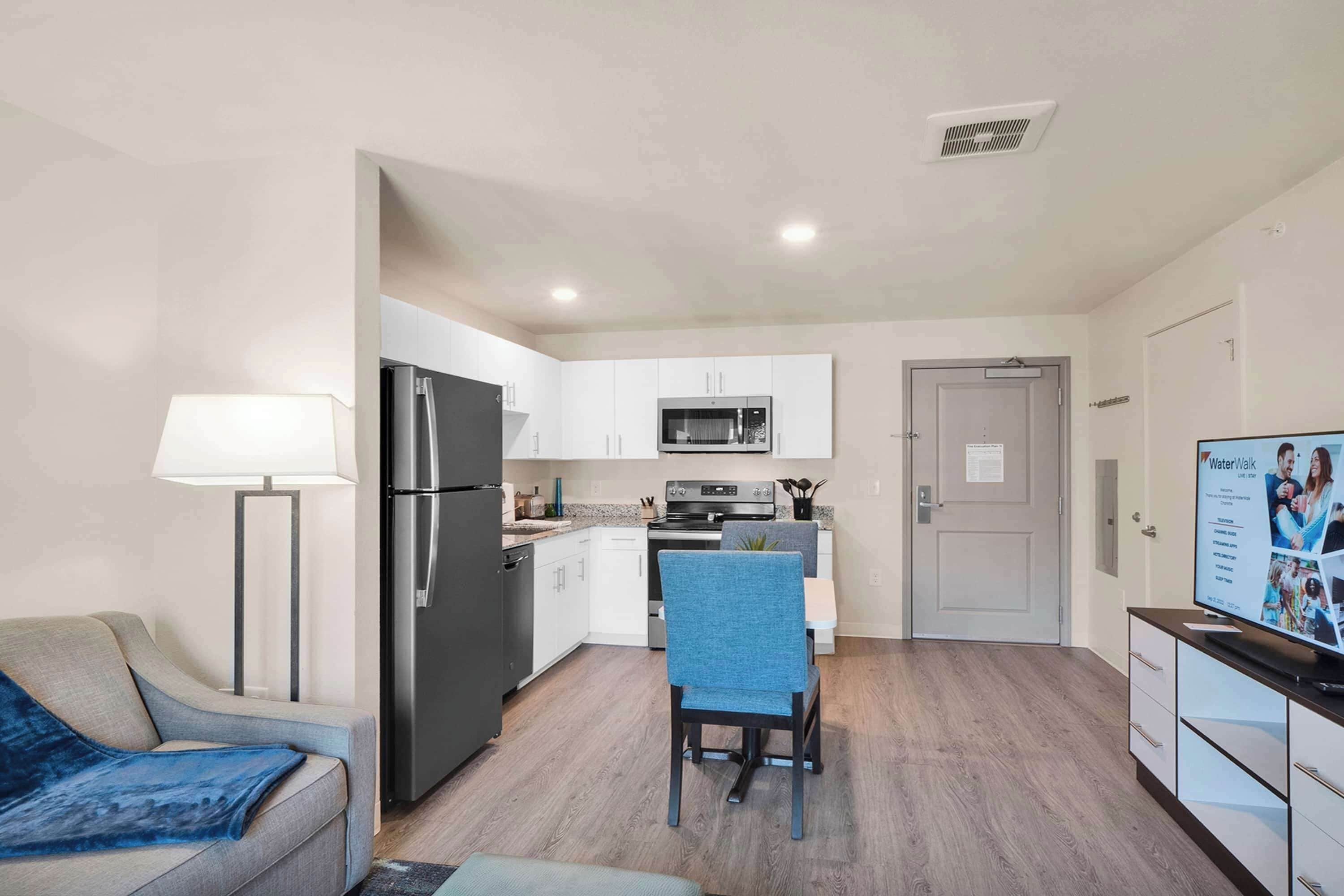 A modern kitchen with white cabinets, a black refrigerator, and a microwave. There is a dining area with a blue chair and a table.