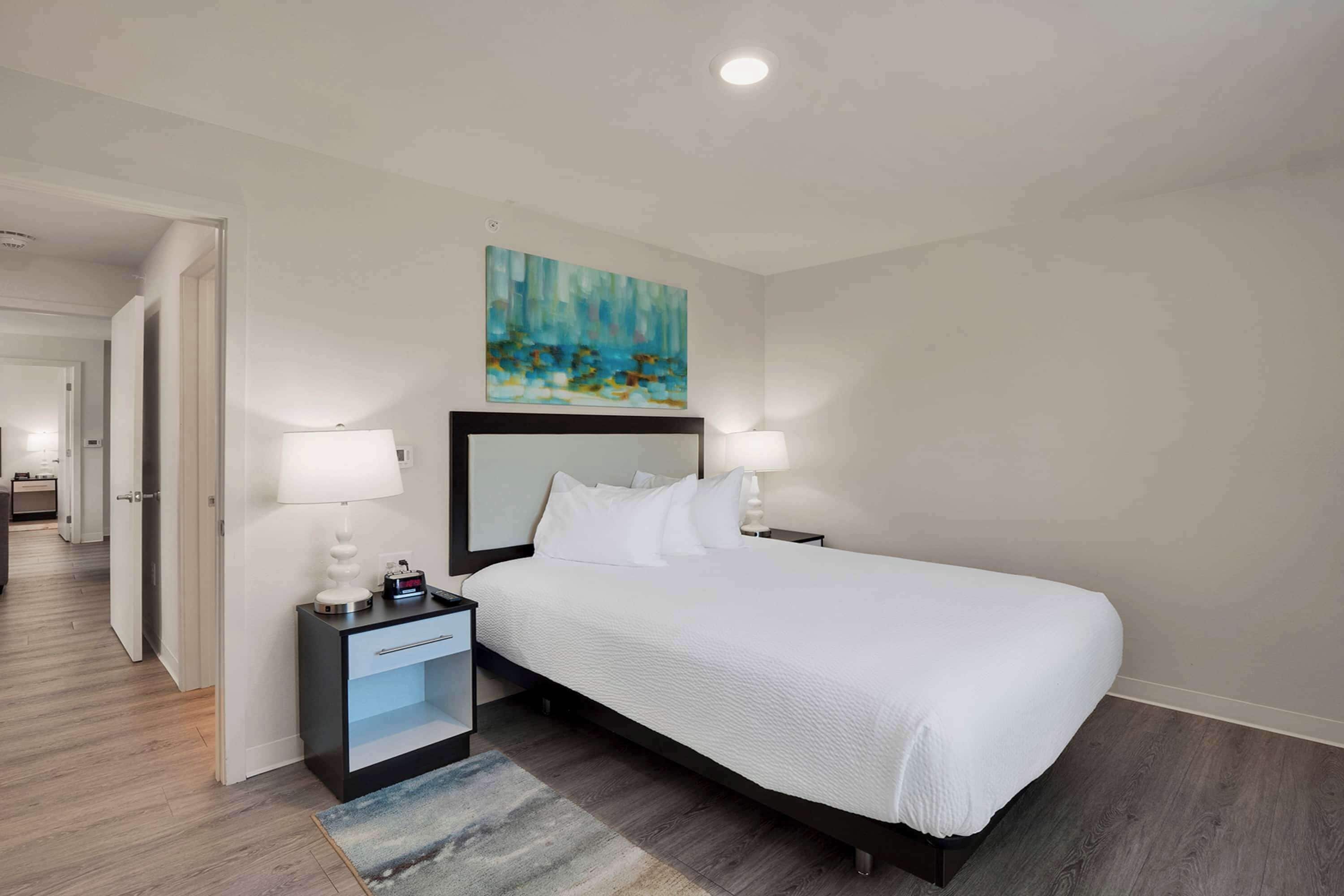 Une chambre d’hôtel moderne avec un grand lit, deux tables de chevet avec des lampes, un tableau au mur et un plancher en bois.