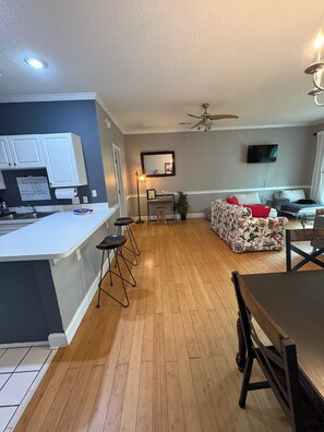 A modern kitchen with a bar area, a dining table, and a living room with a sofa and TV.