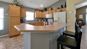 Une cuisine moderne avec des meubles en bois, un îlot central doté d’un comptoir en granit, un four à micro-ondes encastré, un réfrigérateur et un coin repas avec une chaise noire.