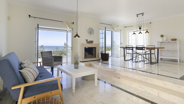 Un vaste espace de vie comprenant un canapé, une table basse et une cheminée. On y trouve également une table à manger avec des chaises, et de grandes fenêtres offrent une vue sur la mer.