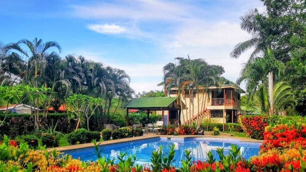 A pool surrounded by vibrant flowers, a two-story building with a green roof, and lush tropical vegetation. - Explore Paradise-Linda Vista cottage (Liberia)