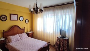 A bedroom with a wooden bed, a nightstand, a lamp, a television on a side table, and a window with curtains.